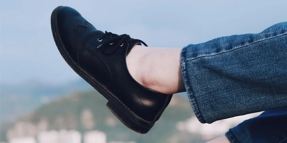 Close-up of a polished black leather shoe with laces, resting against a pair of blue jeans.