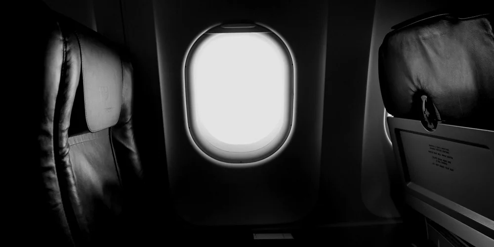 Black-and-white photograph of an airplane cabin interior focusing on leather seats and an overhead window.