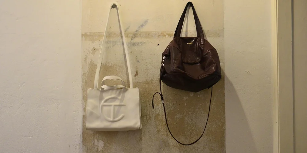 Two leather bags hanging on a wall: a white tote on the left and a dark brown shoulder bag on the right.