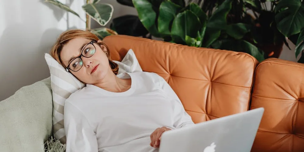 A person wearing a white shirt and glasses relaxes on a brown leather couch, with a laptop on their lap and greenery in the background.