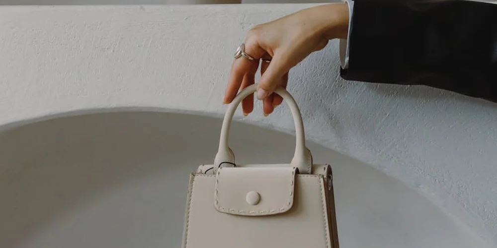 Close-up of a hand lifting a small beige leather handbag with a simple clasp, set against a pale wall.