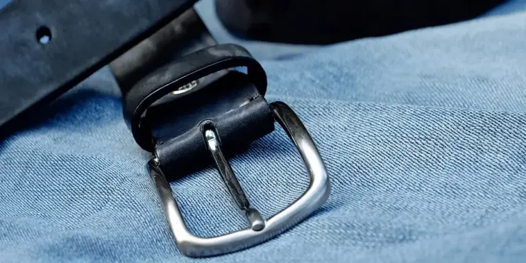 Close-up of a metal belt buckle resting on blue denim fabric, illustrating a leather accessory.