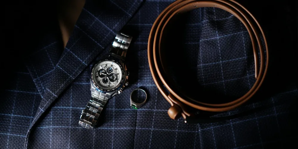 Close-up of a leather belt coiled beside a metal watch on a dark checkered shirt, illustrating belt customization and fit decisions.