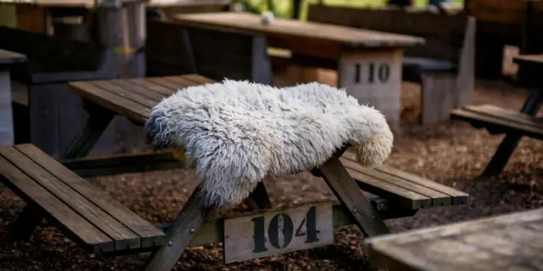 A white fluffy sheepskin draped over a wooden bench in an outdoor setting, with the bench marked '104' in the background.