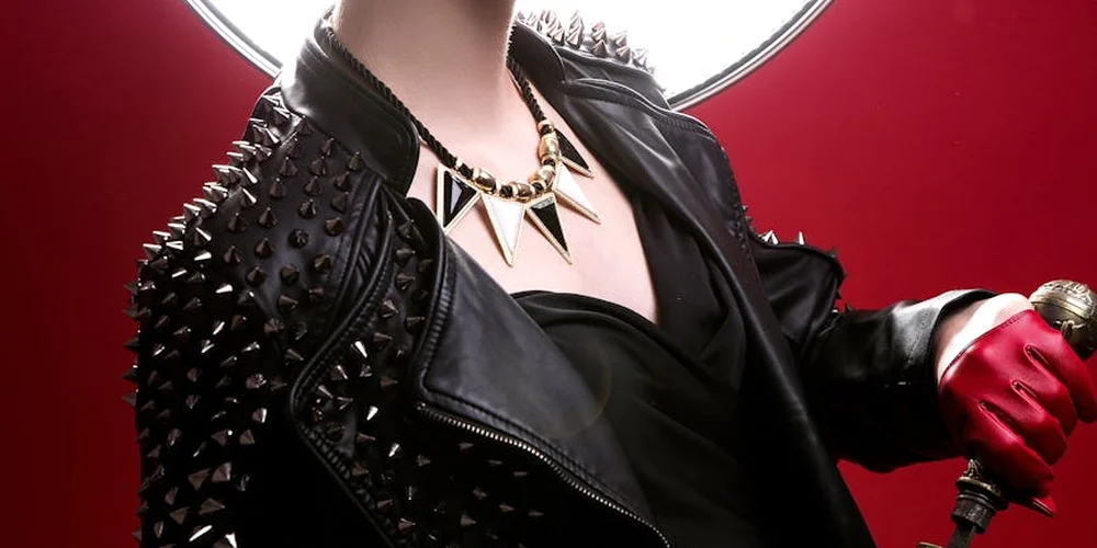 Close-up of a person wearing a black studded leather jacket over a black top, with a chunky necklace, against a red background.