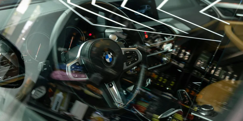 Close-up of a car's steering wheel (BMW) and dashboard, seen through a windshield with reflections.