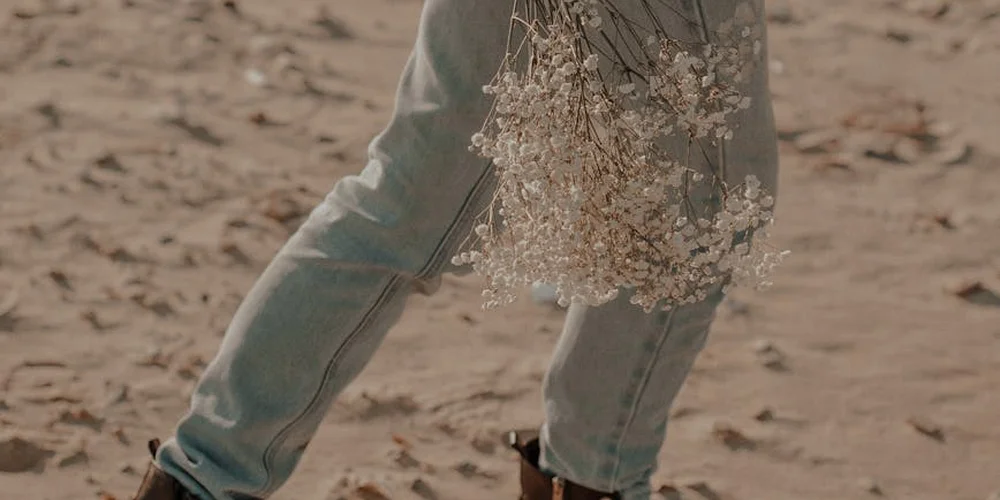 A person stands on sandy ground wearing light-wash jeans and holding dried flowers; the image focuses on textures that help compare leather and suede by touch.