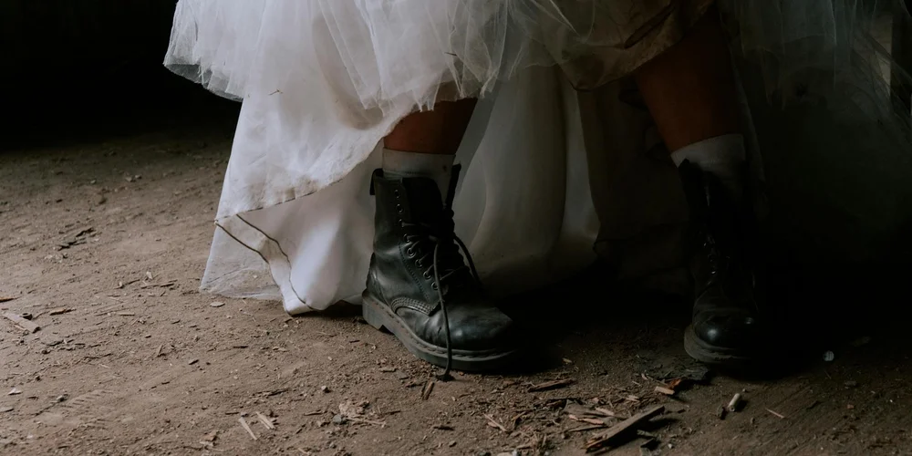 Close-up of black leather boots under a white dress, standing on a dirt floor