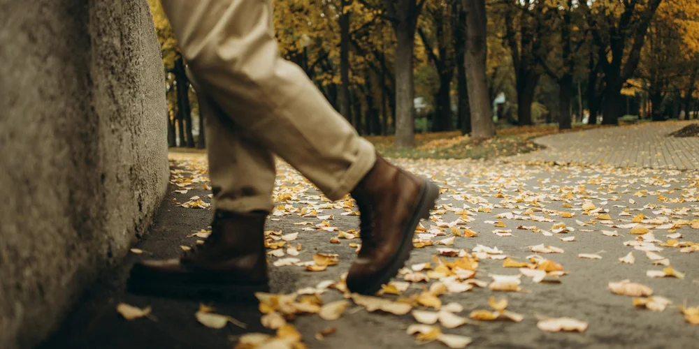 Autumn scene of brown leather boots walking along a leaf-covered sidewalk beside a concrete wall.