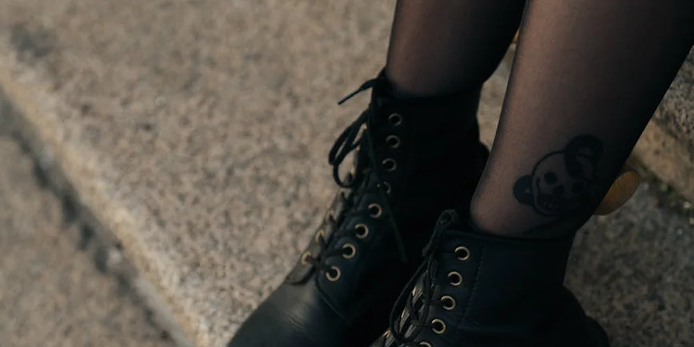Close-up of black lace-up leather boots on a person's legs, showing smooth leather texture and a small ankle tattoo
