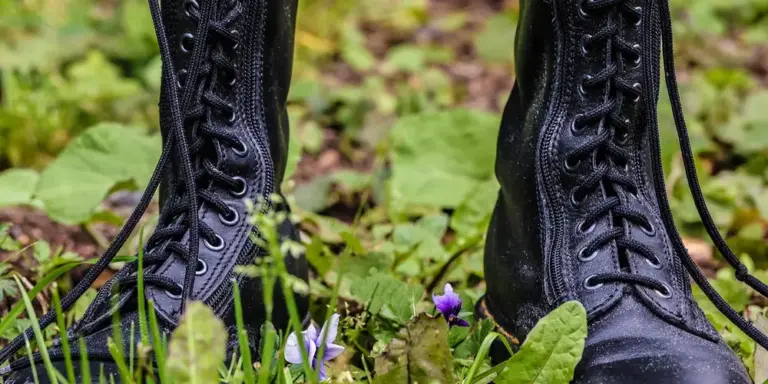 Black lace-up boots standing on grassy ground with green leaves and small purple flowers.