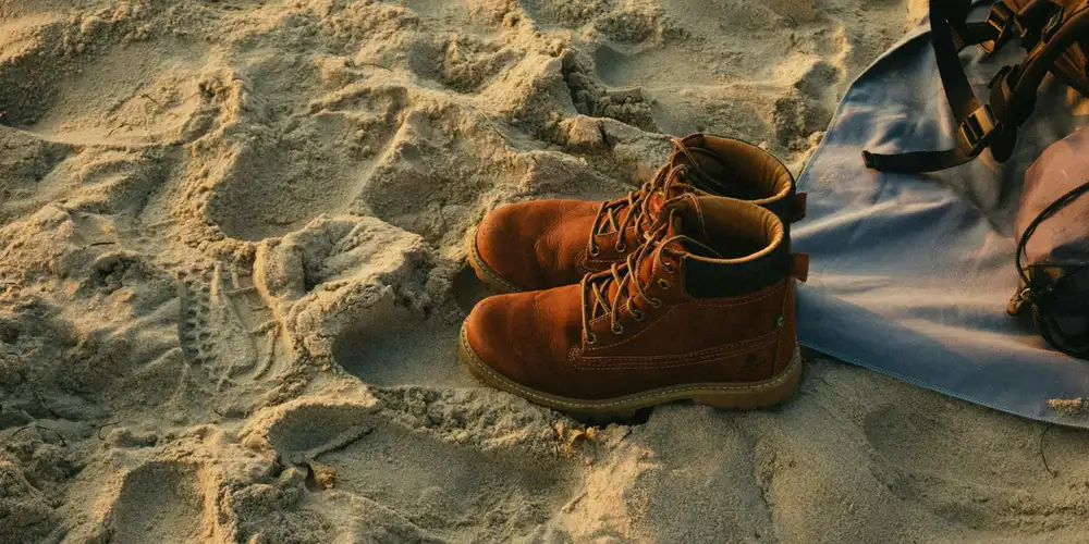 Brown suede boots resting on sandy ground near a blue bag.