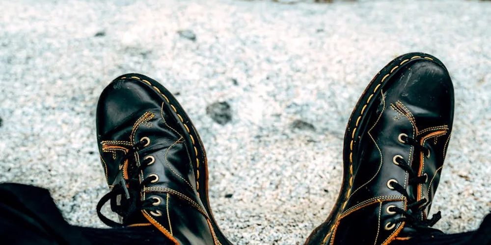 Black leather boots with tan stitching and laces on a textured, light-gray surface, ready for saddle soap care.