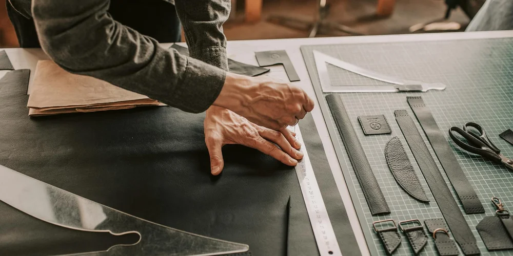 A craftsman’s hands repair a damaged leather piece on a workbench with measuring tools and a cutting mat nearby.