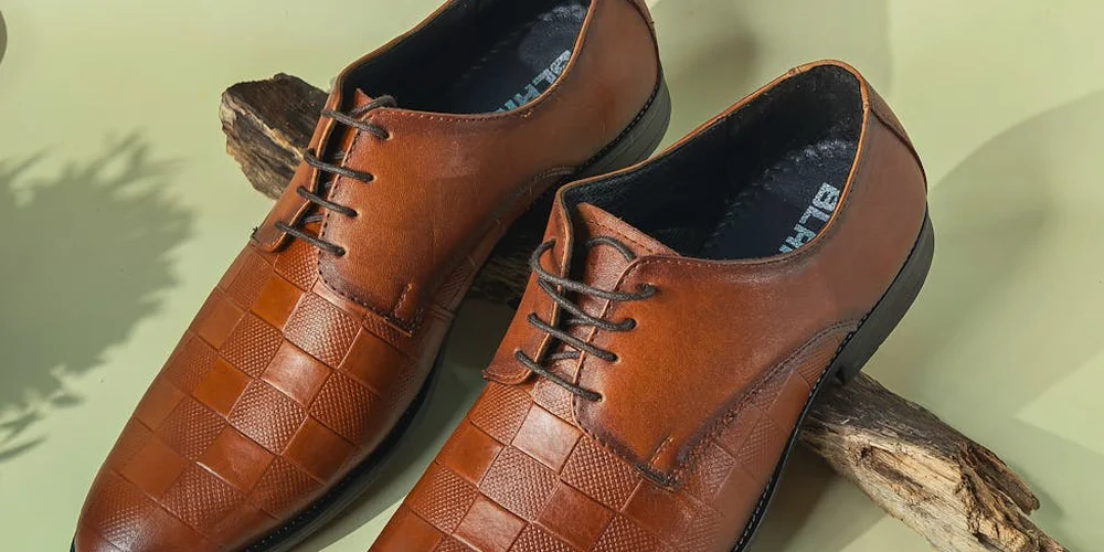 Brown leather dress shoes with a woven toe design on a light green background.