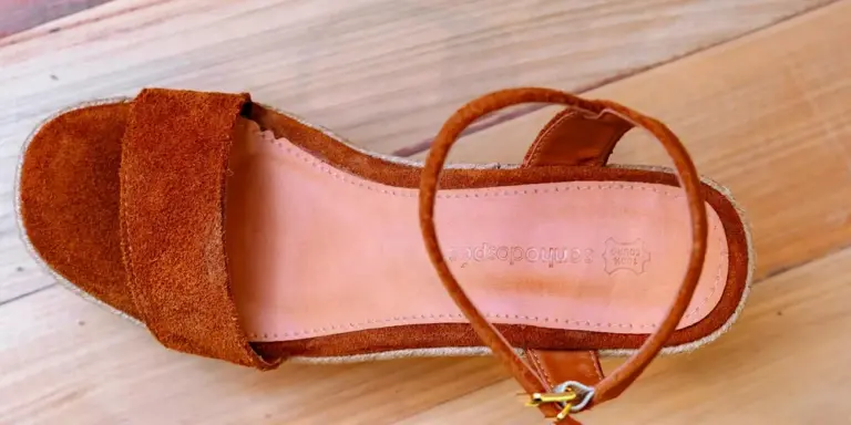 Brown suede open-toe sandal with a strap, placed on a light wooden surface