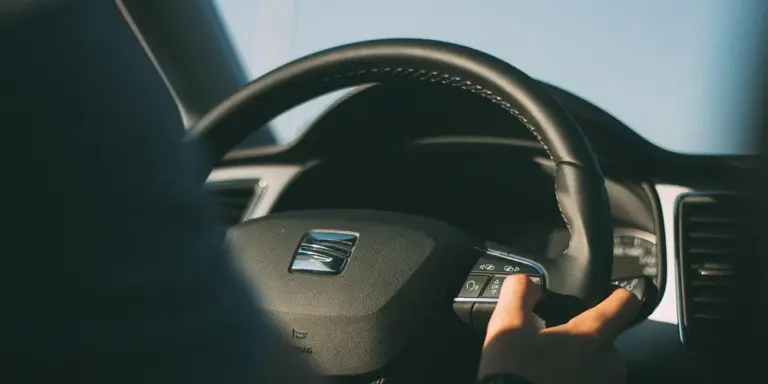 Close-up of a car steering wheel with a hand resting on it inside a leather interior, highlighting the wheel and dashboard area.