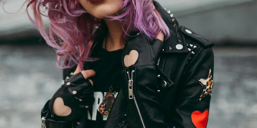 A stylish model with purple hair wearing a black leather jacket featuring heart-shaped cutouts and metal studs.