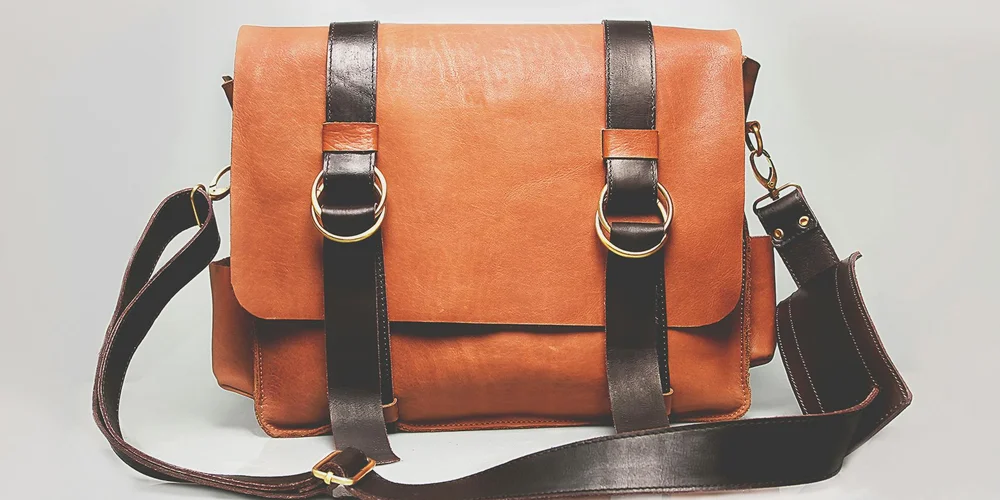 Tan leather messenger bag with dark brown straps and metal buckles on a light background