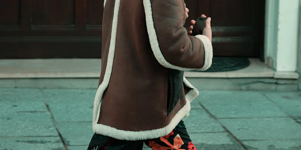 Side view of a person wearing a brown shearling coat with white fur trim, standing on a paved sidewalk.
