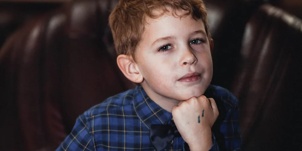 A young boy sits beside a dark leather couch, with a small nail polish smear on his finger.