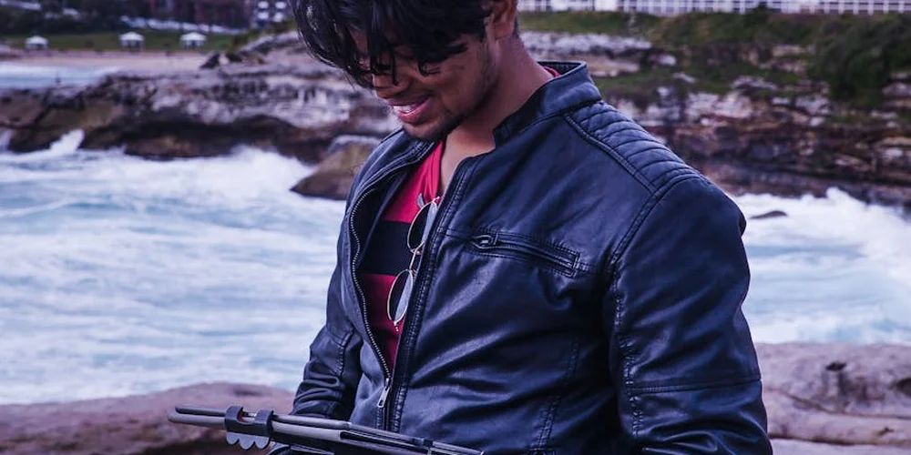 Person wearing a dark leather jacket by the sea, looking down, with sunglasses hanging from the jacket.