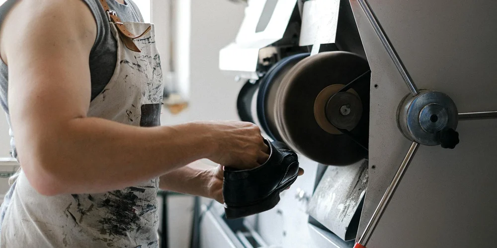 A leatherworker wearing a stained apron polishes a black shoe on a grinding wheel in a workshop.