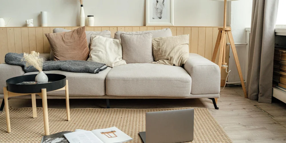 A cozy living room with a light beige sofa, throw pillows, a woven rug, a small coffee table, and a laptop on the floor.