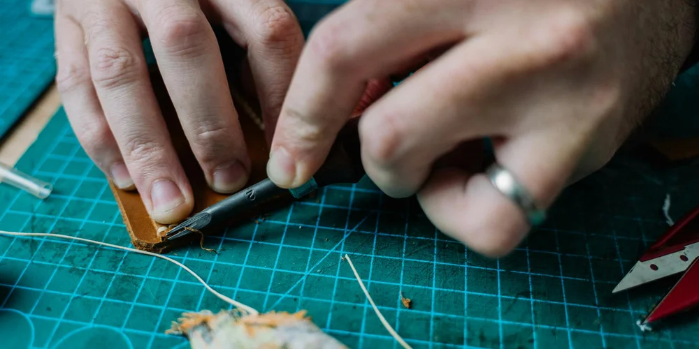 Close-up of hands trimming brown cowhide leather on a green cutting mat with a craft knife.