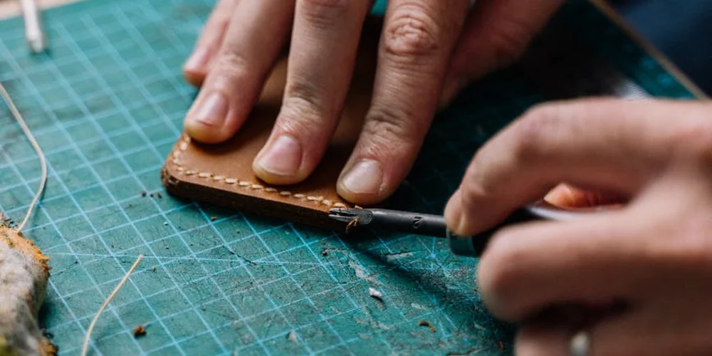 Close-up of hands stitching a piece of cowhide leather on a green cutting mat with a needle and thread.