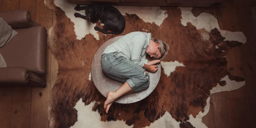A person sits cross-legged on a small round rug placed on top of a curling cowhide rug on a wooden floor, with a dog nearby.