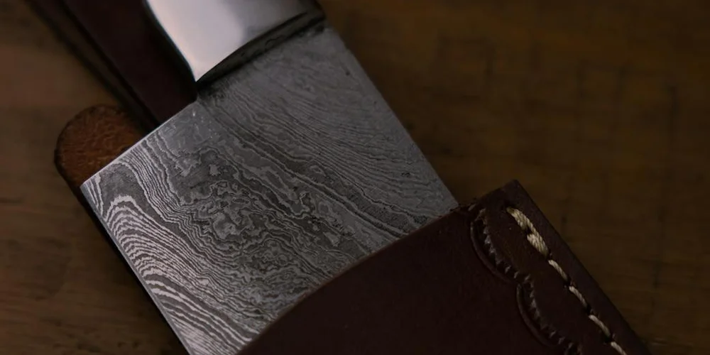 Close-up of a Damascus-pattern knife blade partially sheathed in a dark brown leather sheath on a wooden workbench.