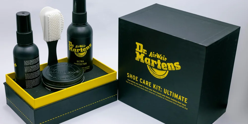Dr. Martens AirWair Shoe Care Kit Ultimate with spray bottle, brush, and other care accessories arranged inside a black and yellow box.