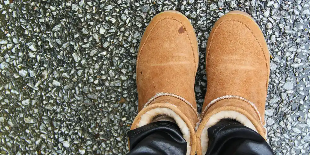Tan suede boots with faux shearling lining standing on a textured, damp pavement.