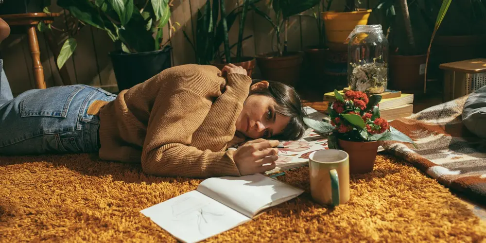 Person lying on a brown faux cowhide rug indoors among plants, with a mug and notebook nearby.