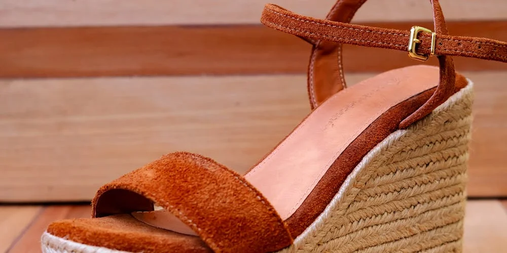 Brown faux suede wedge sandal with an ankle strap resting on a wooden floor.
