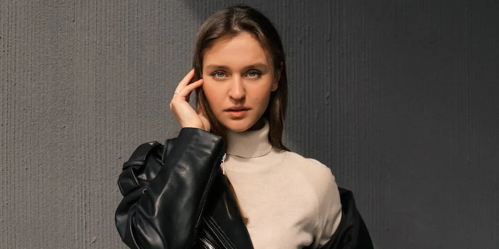 Woman in a beige turtleneck with a black leather jacket draped over her shoulders, ready to inspect the leather.