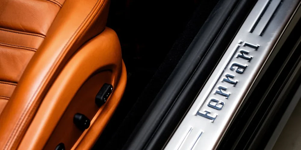 Close-up of orange leather car seat beside a silver Ferrari-branded door sill plate.