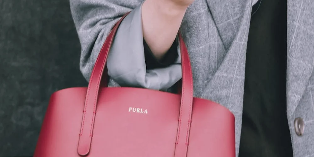Red patent leather Furla handbag with double handles held by a person wearing a gray coat.