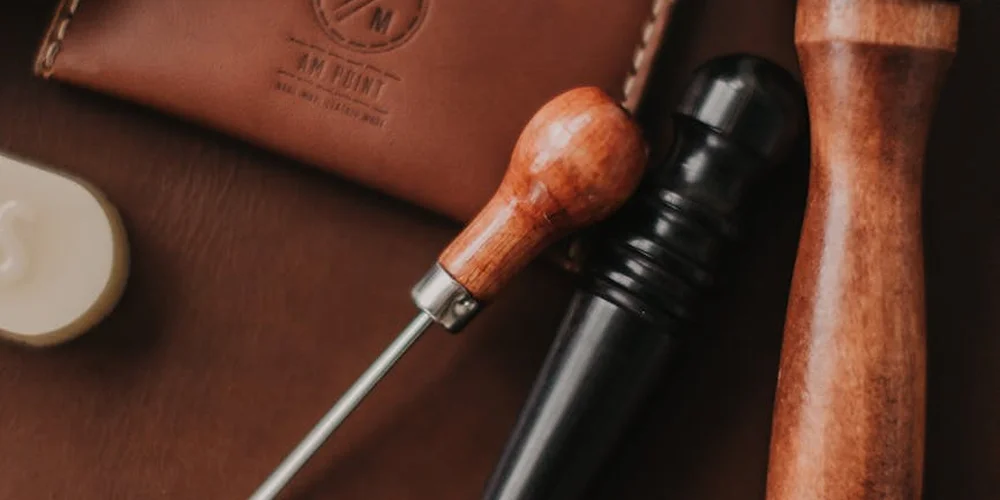 Close-up of leather-care tools on a brown leather surface, including a wooden-handled tool and a leather pouch in the background.