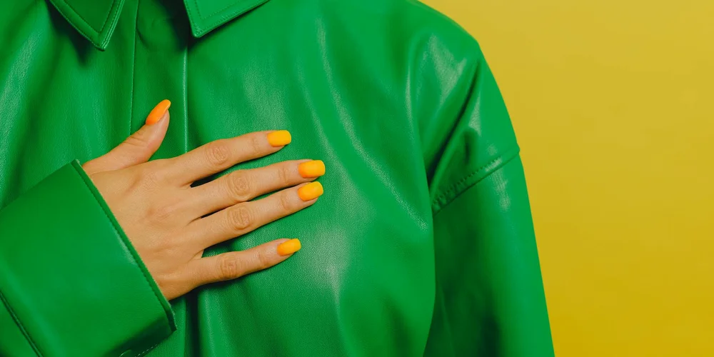 Close-up of a bright green patent leather jacket with a hand resting on the chest against a yellow background.