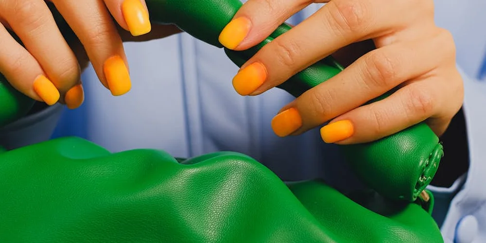 Close-up of hands with bright orange nail polish handling a piece of vivid green leather