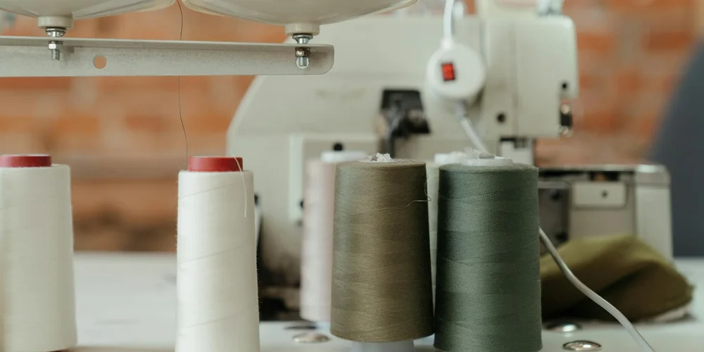 Close-up of spools of thread (white, tan, olive, and brown) on a sewing machine in a leather workshop, suggesting preparation for hand sewing leather projects.