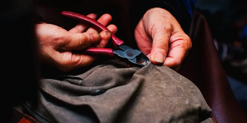 Close-up of hands using red-handled pliers on a piece of leather, illustrating repair work.