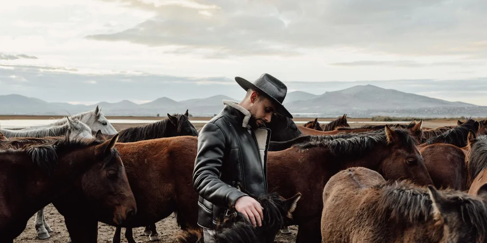 A man wearing a dark leather jacket and a hat stands among horses on a ranch with mountains in the distance.