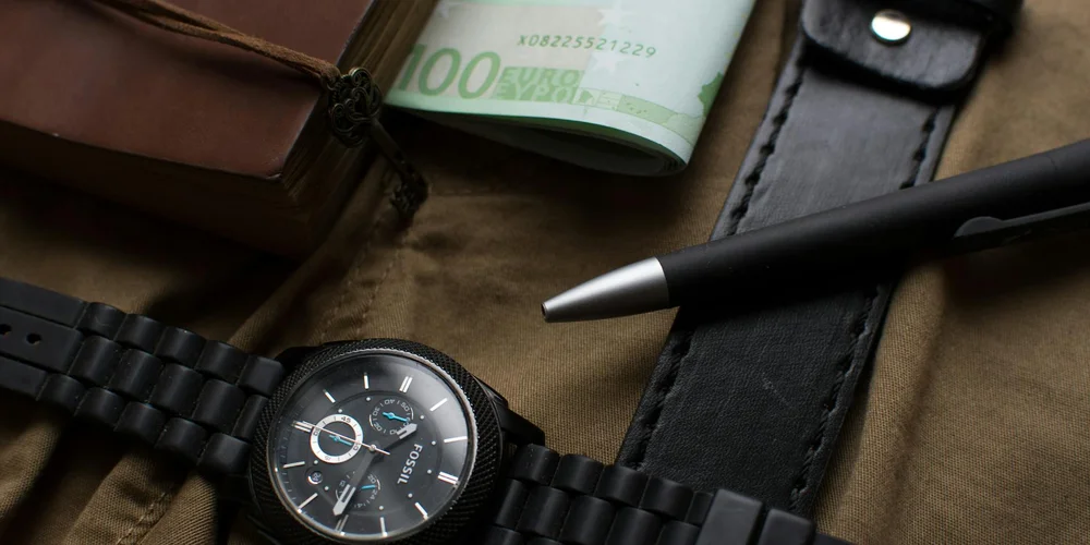 Close-up of a black wristwatch with a leather strap, a rolled hundred-dollar bill, and a black pen on a brown leather surface.