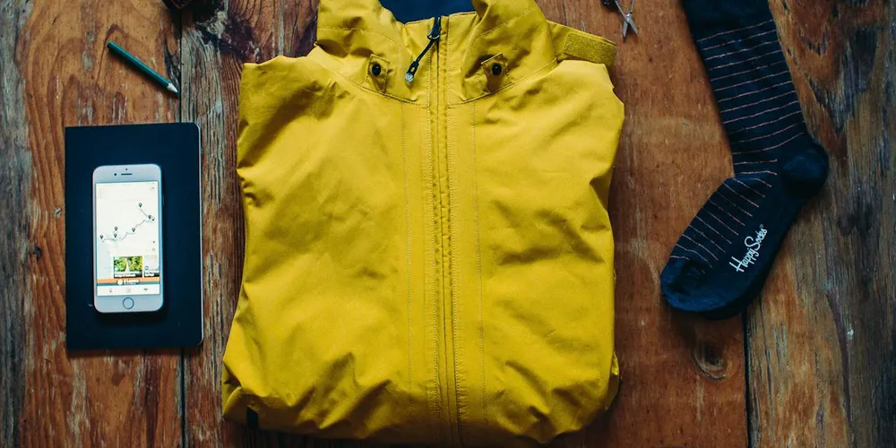 Flat lay of a bright yellow jacket on a wooden floor with a smartphone resting on a small black box and a dark sock nearby.