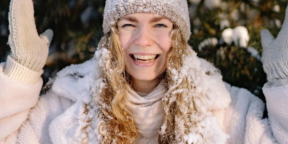 Smiling woman outdoors in the snow, wearing a beige knit hat and light-colored mittens.
