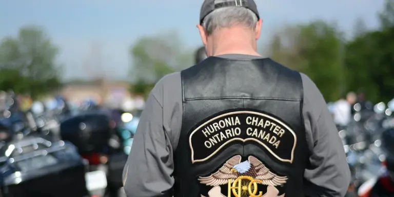 Back view of a person wearing a black leather vest with a Huronia Chapter Ontario Canada patch, standing among motorcycles.