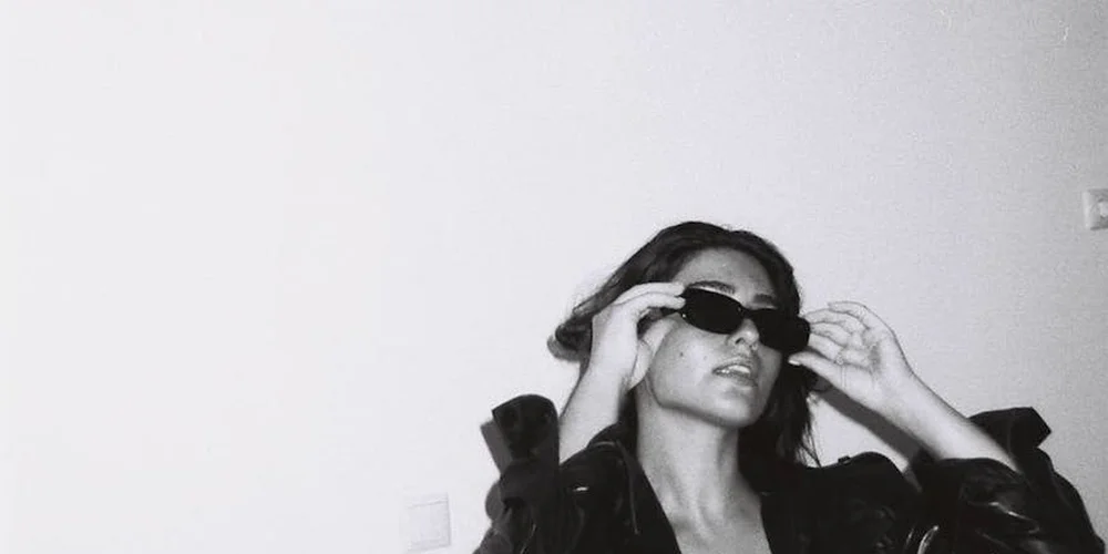 A person wearing dark sunglasses adjusts them in a black-and-white portrait.